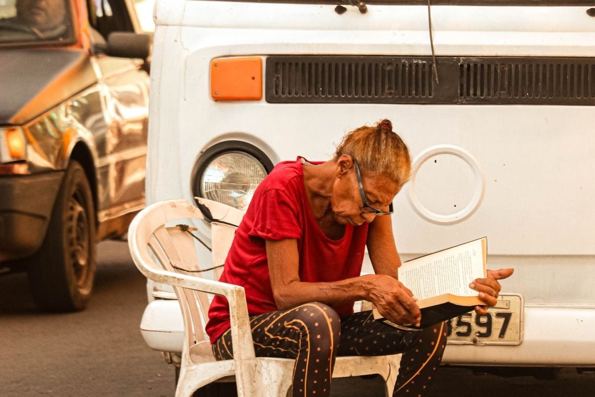 Senhora sentada numa cadeira na calçada, lendo, numa rua de cidade