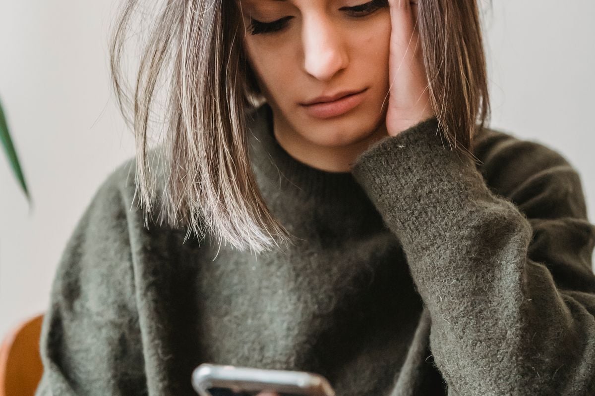 Jovem sentada em casa lendo uma mensagem no celular, com ar preocupado
