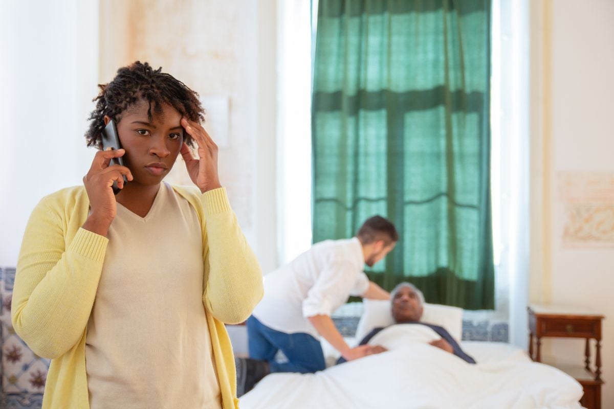 Mulher ao telefone, aflita, enquanto um cuidador atende uma pessoa idosa na cama